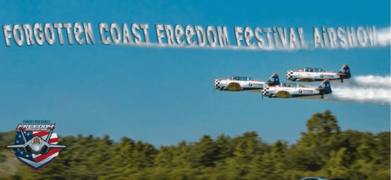 Promotional banner for the Forgotten Coast Freedom Festival Air Show. Against a bright blue sky, three small prop planes fly in formation from right to left, leaving a white smoke trail that forms the words ‘FORGOTTEN COAST FREEDOM FESTIVAL AIR SHOW’ across the top. A line of trees and an airfield runway run along the bottom, with two sports cars on the far right. The event logo—featuring a jet over an American-flag shield—appears in the lower-left corner.
